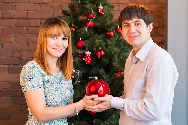 Pareja joven romántica con bola de Navidad cerca del árbol de Navidad