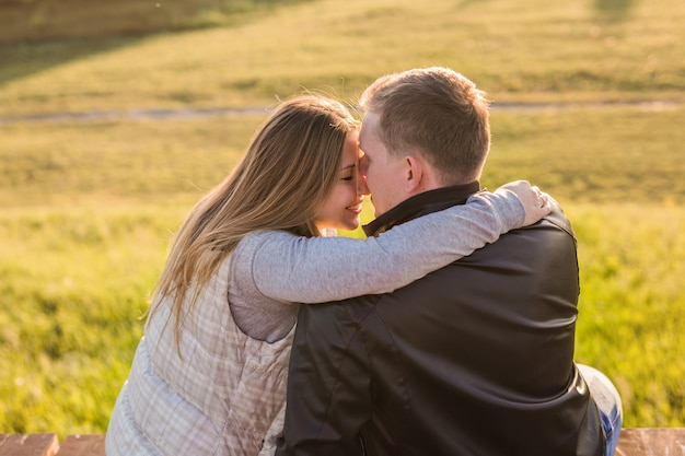 Pareja joven romántica disfrutando de una cita sentados en un estrecho abrazo en un banco del parque con vistas a un lago, vista desde atrás