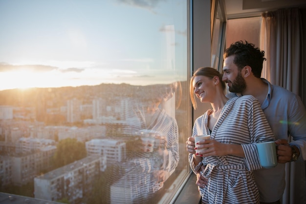 pareja joven romántica y feliz disfrutando del café de la tarde y el hermoso paisaje de la puesta de sol de la ciudad mientras está de pie junto a la ventana
