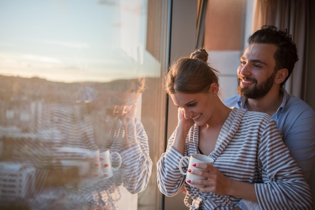 pareja joven romántica y feliz disfrutando del café de la tarde y el hermoso paisaje de la puesta de sol de la ciudad mientras está de pie junto a la ventana