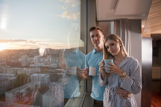 pareja joven romántica y feliz disfrutando del café de la tarde y el hermoso paisaje de la puesta de sol de la ciudad mientras está de pie junto a la ventana