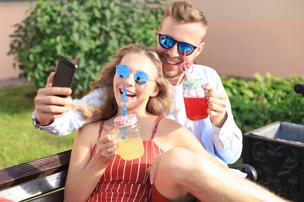 Pareja joven romántica en ropa de verano sonriendo y tomando selfie mientras está sentado en un banco en las calles de la ciudad.