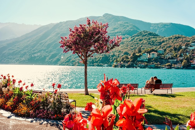 Pareja joven romántica sentada en un banco en el complejo turístico de lujo de Ascona en el lago Maggiore en el cantón de Ticino en Suiza en verano. La gente viaja por la calle al aire libre. Vacaciones caras de personas familiares.