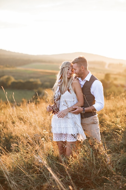 Pareja joven en ropa rústica boho elegante abrazando, de pie en el campo al atardecer