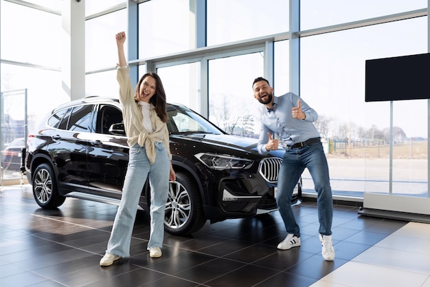 Pareja joven satisfecha bailando junto a un auto nuevo comprado en un concesionario de autos