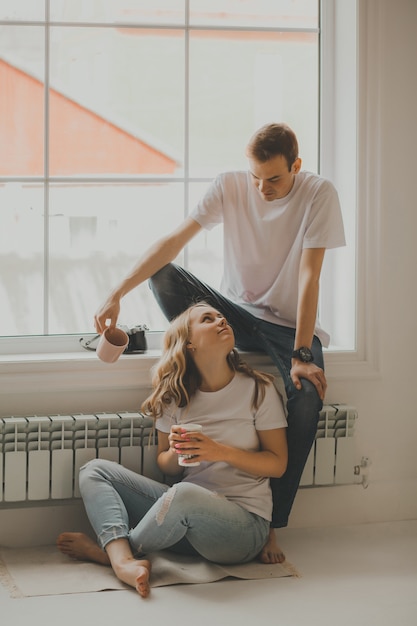 Foto una pareja joven está sentada cerca de la ventana grande, descansando y tomando café en tazas después de una sesión de fotos, una cámara retro está en el alféizar de la ventana.