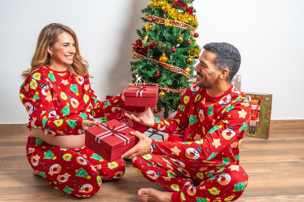 Pareja joven sentada dando regalos de Navidad junto al árbol de Navidad Familia con mujer embarazada