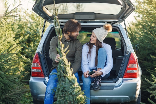 Pareja joven sentada en el maletero de su SUV sosteniendo un árbol de Navidad recién cortado en una plantación