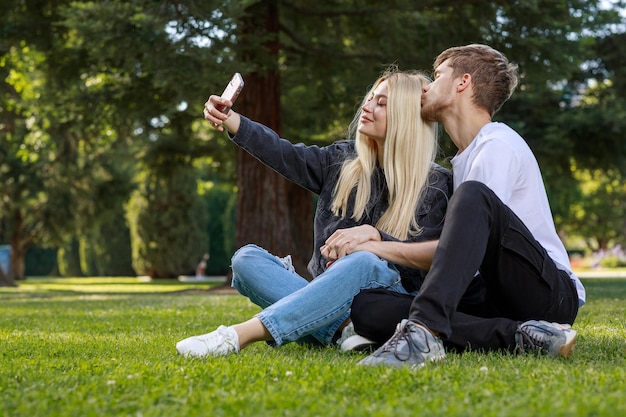 Pareja joven sentada en el parque en el césped y tomando selfie