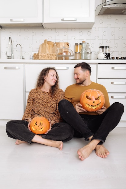 Pareja joven sentada en el piso de la cocina y sosteniendo calabazas de Halloween