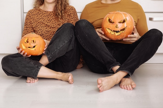 Pareja joven sentada en el piso de la cocina y sosteniendo calabazas de Halloween