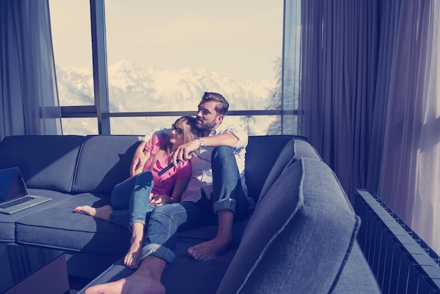 Una pareja joven sentada en un sofá cerca de la ventana viendo la televisión juntos en una casa de lujo