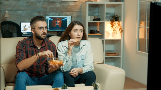 Pareja joven sentada en el sofá y viendo la televisión disfrutando de sus patatas fritas. Pareja mirando tensa en la televisión.