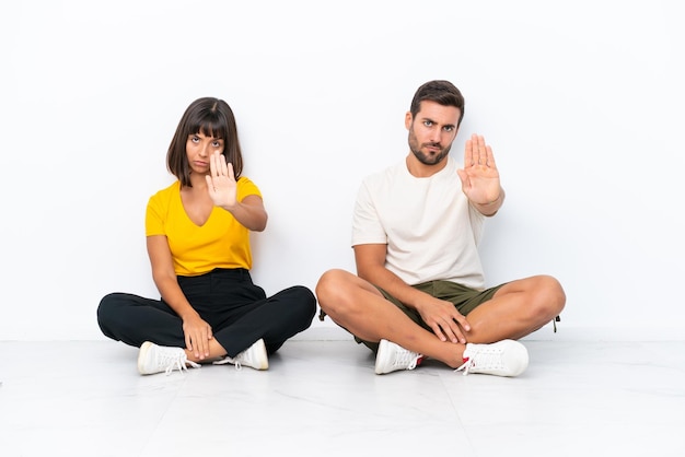 Una pareja joven sentada en el suelo aislada de fondo blanco haciendo un gesto de parada negando una situación que piensa mal