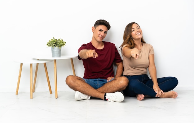 Pareja joven sentada en el suelo en el frente blanco apuntando con expresión feliz