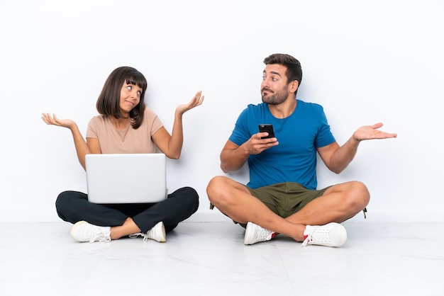 Foto pareja joven sentada en el suelo sosteniendo pc y teléfono móvil aislado sobre fondo blanco haciendo gestos sin importancia mientras levanta los hombros