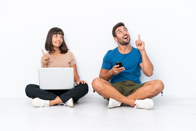 Foto pareja joven sentada en el suelo sosteniendo pc y teléfono móvil aislado sobre fondo blanco señalando con el dedo índice una gran idea