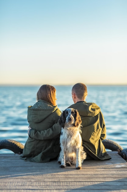 Una pareja joven se sienta con un perro en un muelle junto al mar al atardecer