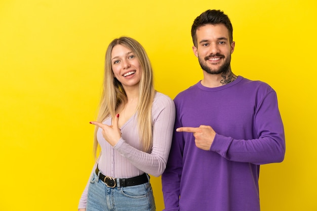 Pareja joven sobre fondo amarillo aislado apuntando con el dedo hacia un lado y presentando un producto