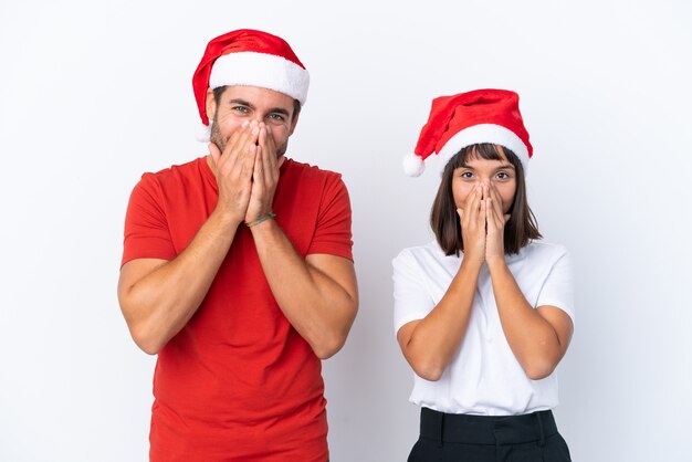 Pareja joven con sombrero de navidad aislado sobre fondo blanco sonriendo mucho mientras cubre la boca