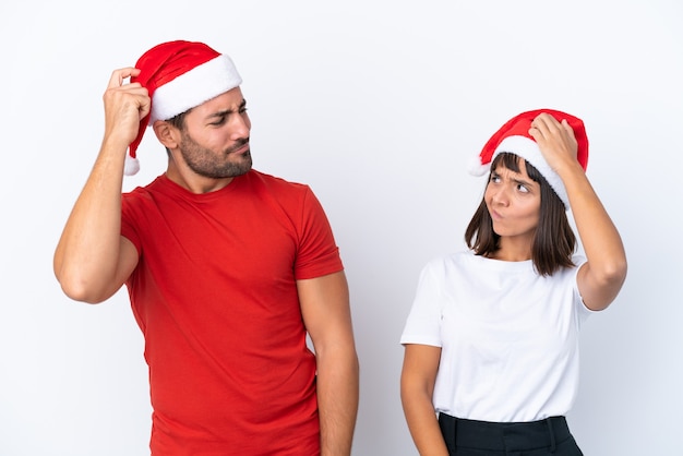 Pareja joven con sombrero de navidad aislado sobre fondo blanco teniendo dudas mientras se rasca la cabeza