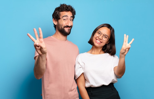 Pareja joven sonriendo y mirando amigable, mostrando el número tres o tercero con la mano hacia adelante, contando hacia atrás