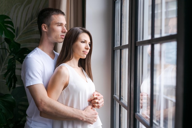 Pareja joven sonriente abrazándose y de pie cerca de la ventana mirando