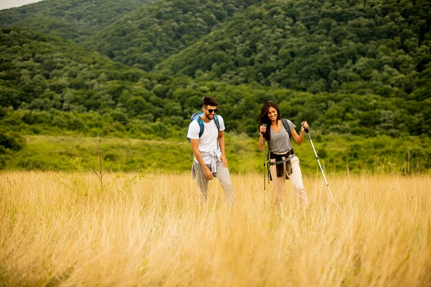 Pareja joven sonriente caminando con mochilas sobre colinas verdes