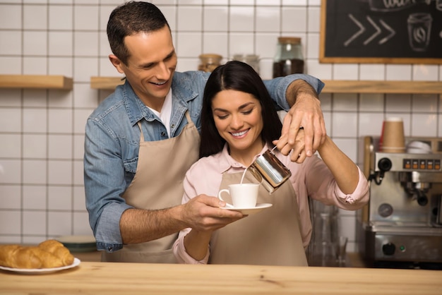 Pareja joven sonriente en delantales preparando café juntos en la cafetería