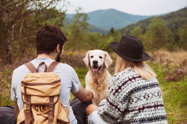 Pareja joven con un sonriente perro golden retriever sentado en la hierba en el valle de las montañas de verano. Concepto de viajes de mascotas, senderismo y actividades de fin de semana. Mujer de cabello rubio con sombrero y hombre con mochila.