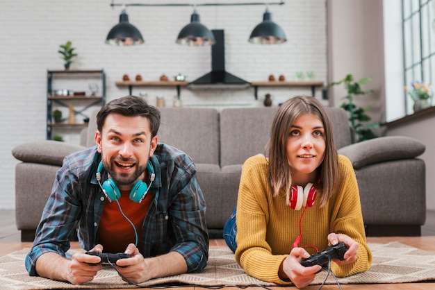 Pareja joven sonriente que miente en el piso que juega al videojuego con la palanca de mando en casa