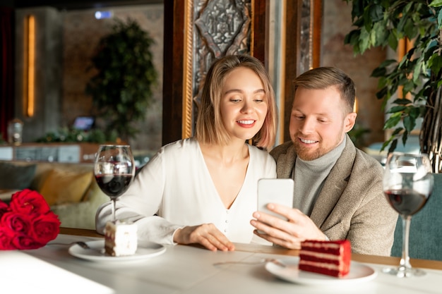 Pareja joven sonriente viendo algo en el teléfono inteligente mientras está sentado junto a la mesa servida en un elegante restaurante y tomando vino y pasteles