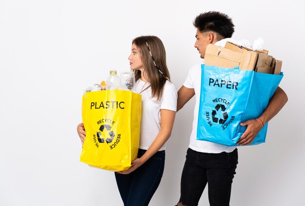 Foto pareja joven sosteniendo una bolsa llena de plástico y papel en blanco extendiendo las manos hacia un lado para invitar a venir