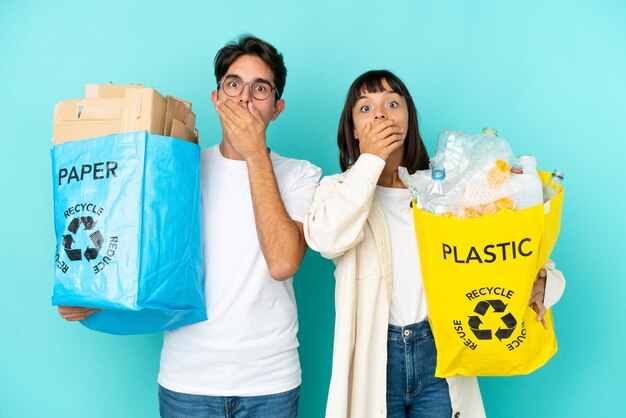 Pareja joven sosteniendo una bolsa llena de plástico y papel para reciclar aislada en un fondo azul cubriendo la boca con las manos por decir algo inapropiado