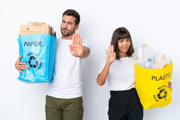 Una pareja joven sosteniendo una bolsa llena de plástico y papel para reciclar aislada de fondo blanco haciendo un gesto de parada negando una situación que piensa mal