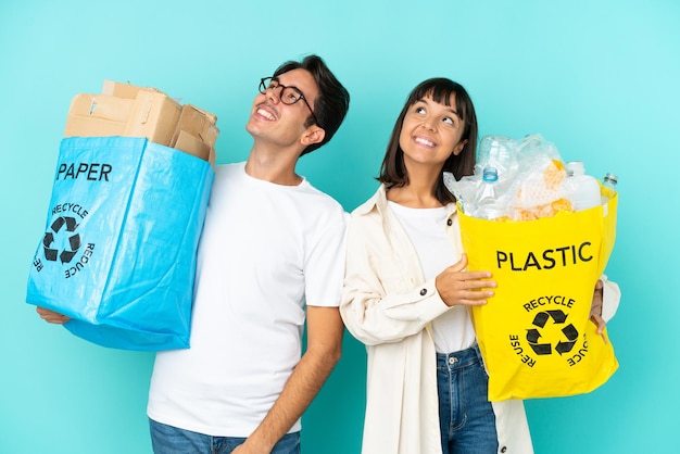 Pareja joven sosteniendo una bolsa llena de plástico y papel para reciclar aislado sobre fondo azul mirando hacia arriba mientras sonríe