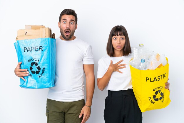 Pareja joven sosteniendo una bolsa llena de plástico y papel para reciclar aislado sobre fondo blanco con expresión facial sorprendida y conmocionada