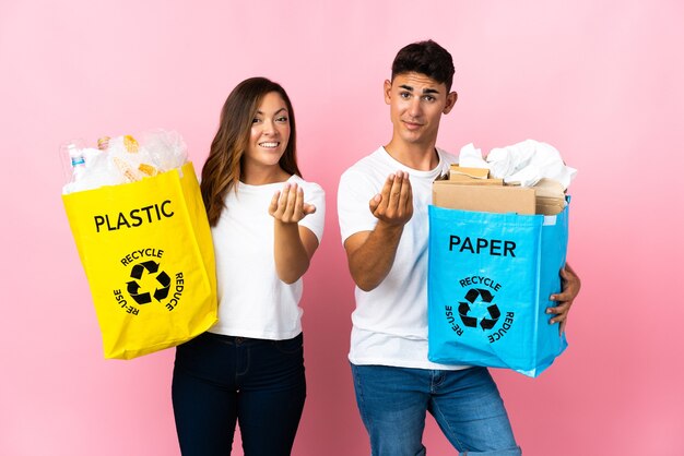 Pareja joven sosteniendo una bolsa llena de plástico y papel en rosa invitando a venir con la mano.