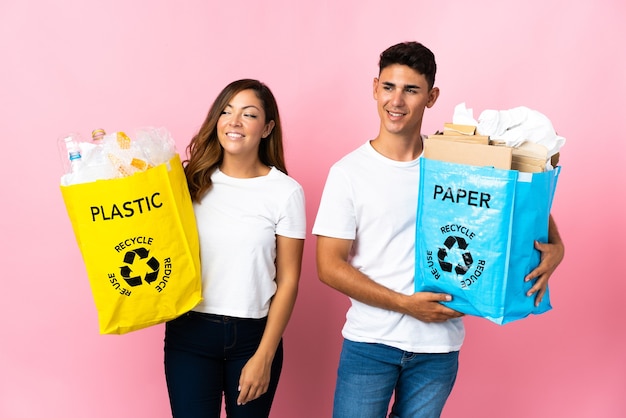 Pareja joven sosteniendo una bolsa llena de plástico y papel en rosa mirando hacia un lado y sonriendo