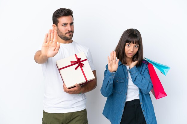 Pareja joven sosteniendo bolsas de la compra y presente aislado sobre fondo blanco haciendo gesto de parada negando una situación que piensa mal