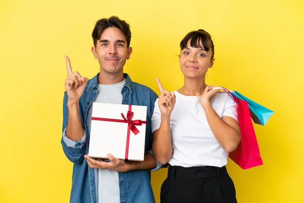 Pareja joven sosteniendo bolsas de compras y presente aislado sobre fondo amarillo mostrando y levantando un dedo en señal de lo mejor