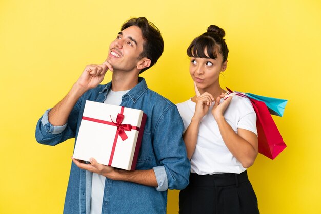 Pareja joven sosteniendo bolsas de compras y presente aislado sobre fondo amarillo pensando en una idea mientras mira hacia arriba