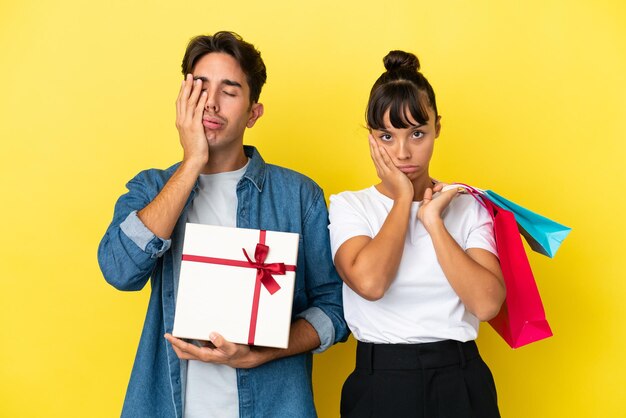 Foto pareja joven sosteniendo bolsas de compras y presente aislado sobre fondo amarillo con sorpresa y expresión facial conmocionada