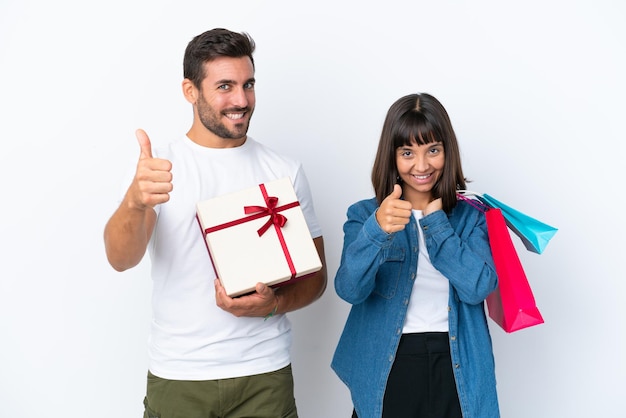 Pareja joven sosteniendo bolsas de compras y presente aislado sobre fondo blanco dando un gesto de aprobación porque algo bueno ha sucedido