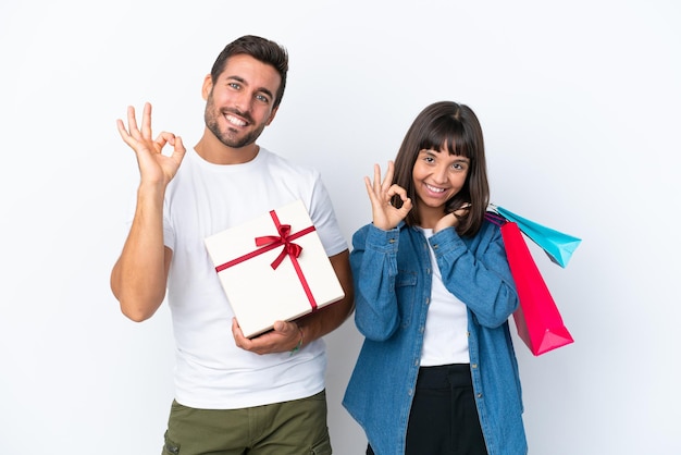 Pareja joven sosteniendo bolsas de compras y presente aislado sobre fondo blanco mostrando un signo de ok con los dedos