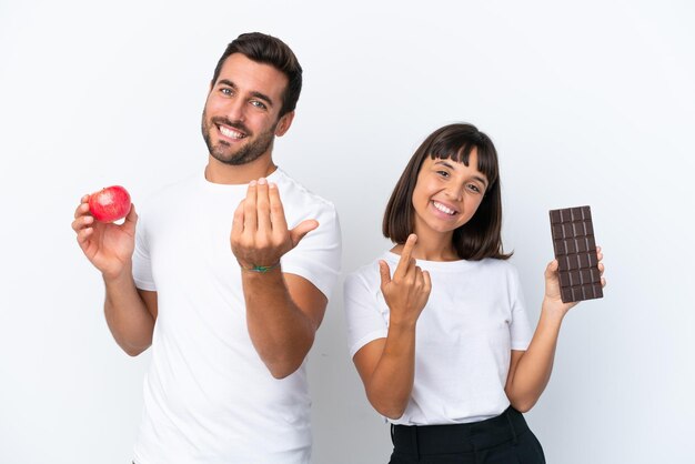 Pareja joven sosteniendo chocolate y manzana aislado sobre fondo blanco invitando a venir con la mano Feliz de que hayas venido