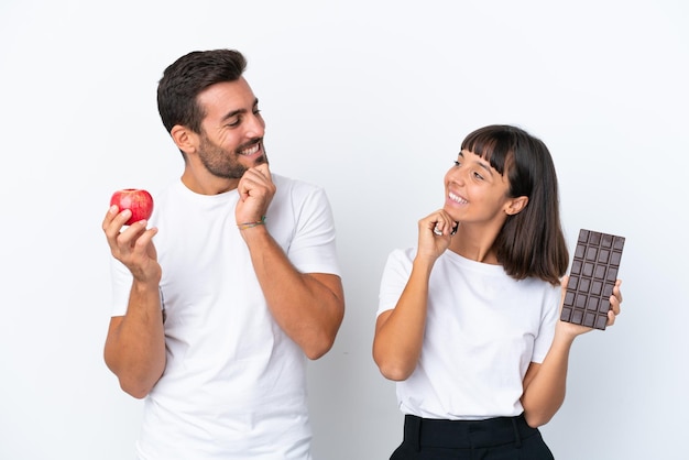 Pareja joven sosteniendo chocolate y manzana aislado sobre fondo blanco mirando el uno al otro