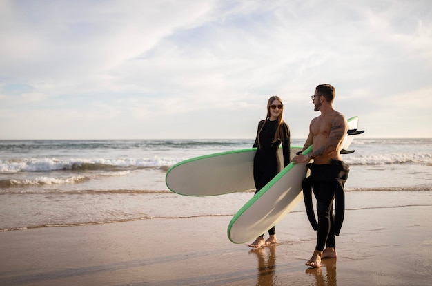 Una pareja joven de surfistas camina con tablas de surf en la playa al atardecer