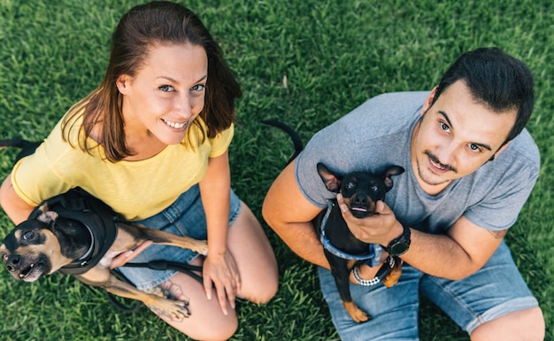 Pareja joven con sus perros sentados en el césped y mirando hacia arriba en el parque. Vista superior de la pareja sentada en la hierba.