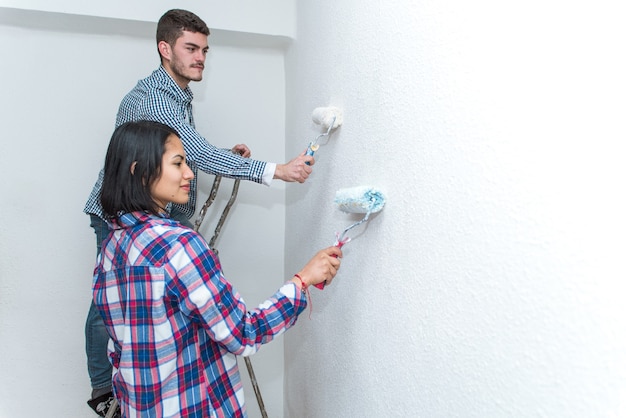 Pareja joven trabajando juntos en la renovación de su nuevo apartamento. Hombre y mujer pintando la pared blanca con rodillos.